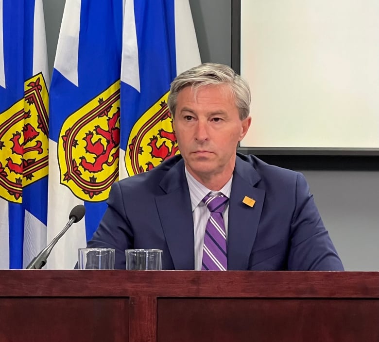 A man wearing a suit and tie sits at a desk with a microphone in front of Nova Scotia flags.