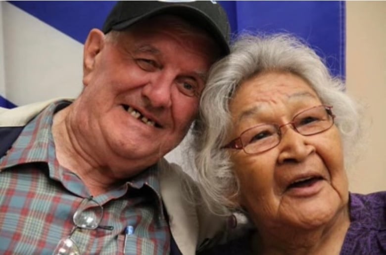 An older man wearing a ball cap poses with a old woman wearing glass, holding each other close while smiling.