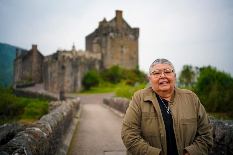 An older woman with silver hair and glasses poses in front of a castle.