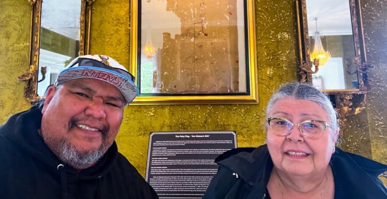 an older women with glasses and silver hair poses beside a man with a hat and sunglasses, in front of an old flag.