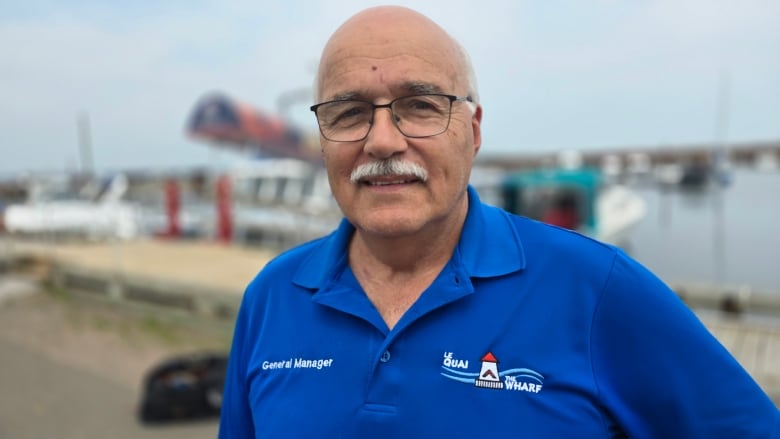 A bespectacled man with a white mustache and wearing a blue shirt stands on a dock.