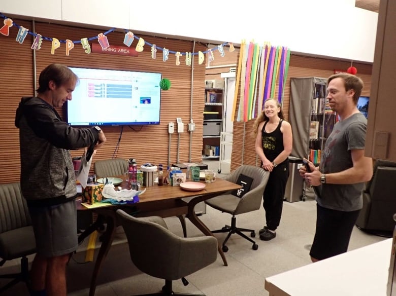 A man opens presents as two people watch and smile. Birthday streamers hang from the wall