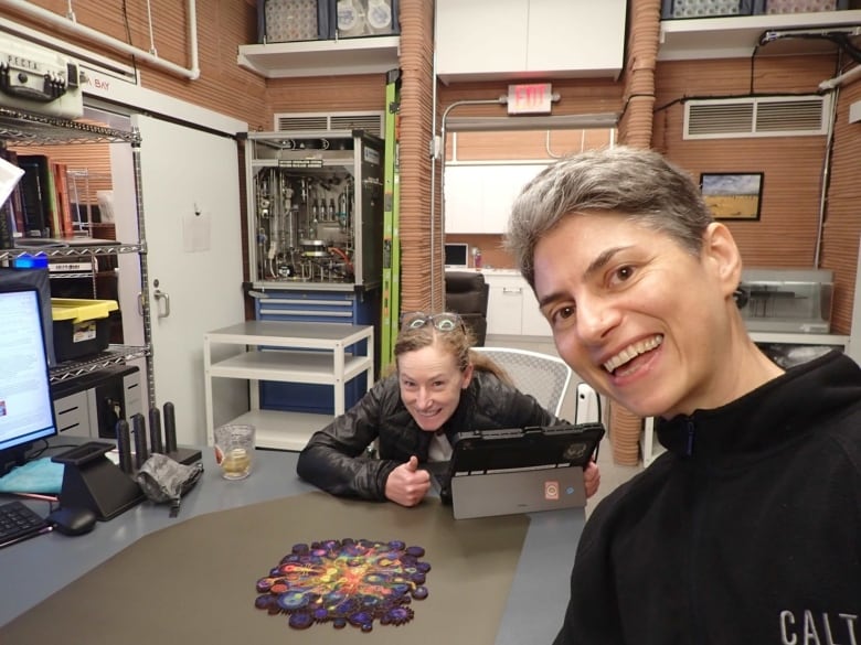 Two smiling women take a selfie in a wood-panneled room full of tech equipment. There's a puzzle or a board game on the table between them