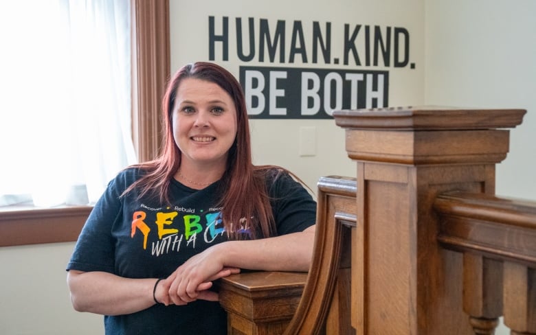 A person is seen leaning at the top of a wooden bannister, smiling. Behind them is a sign on the wall that says, "Human. Kind. Be Both."