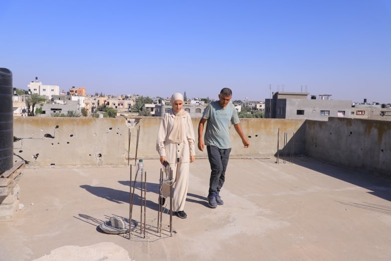 Two people walk on a flat roof of a building, with other buildings in the background.