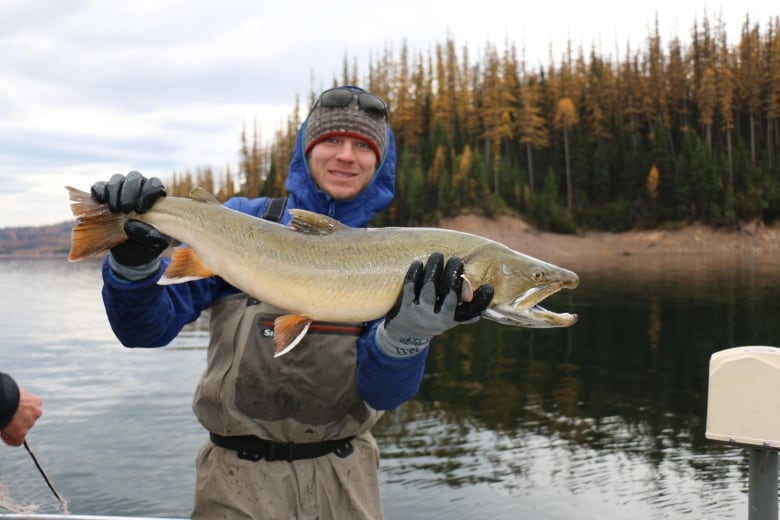 Man in a grew and blue jacket holds a fish in front of a lake. 