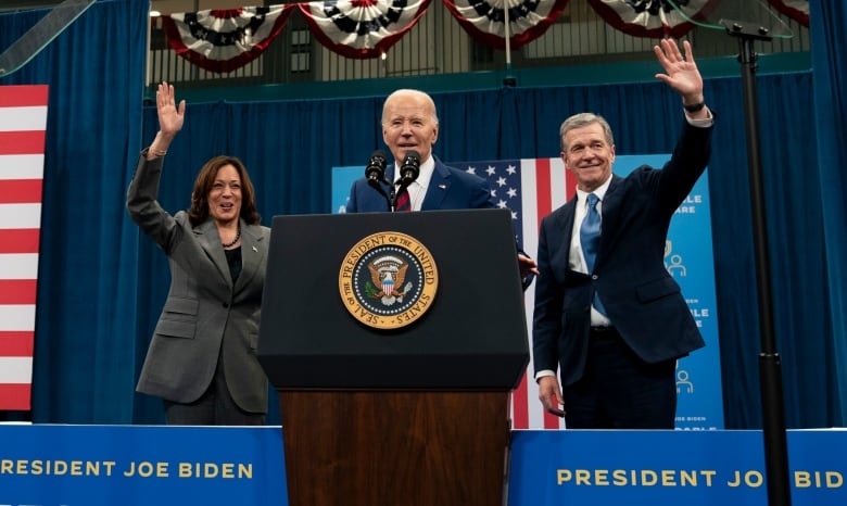 A waving man and woman flank a second man standing at a podium. 