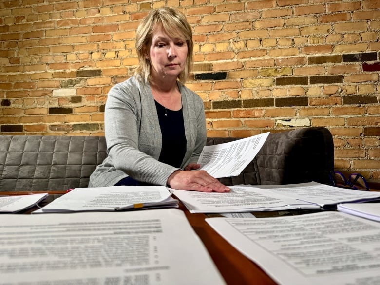 A woman wearing a grey sweater is seated on a couch with numerous documents on a table in front of her.