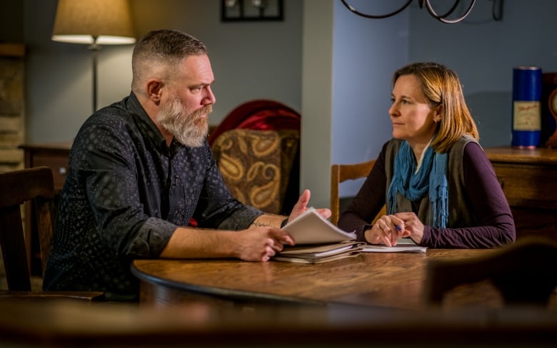 Man with beard talks to woman at a table.