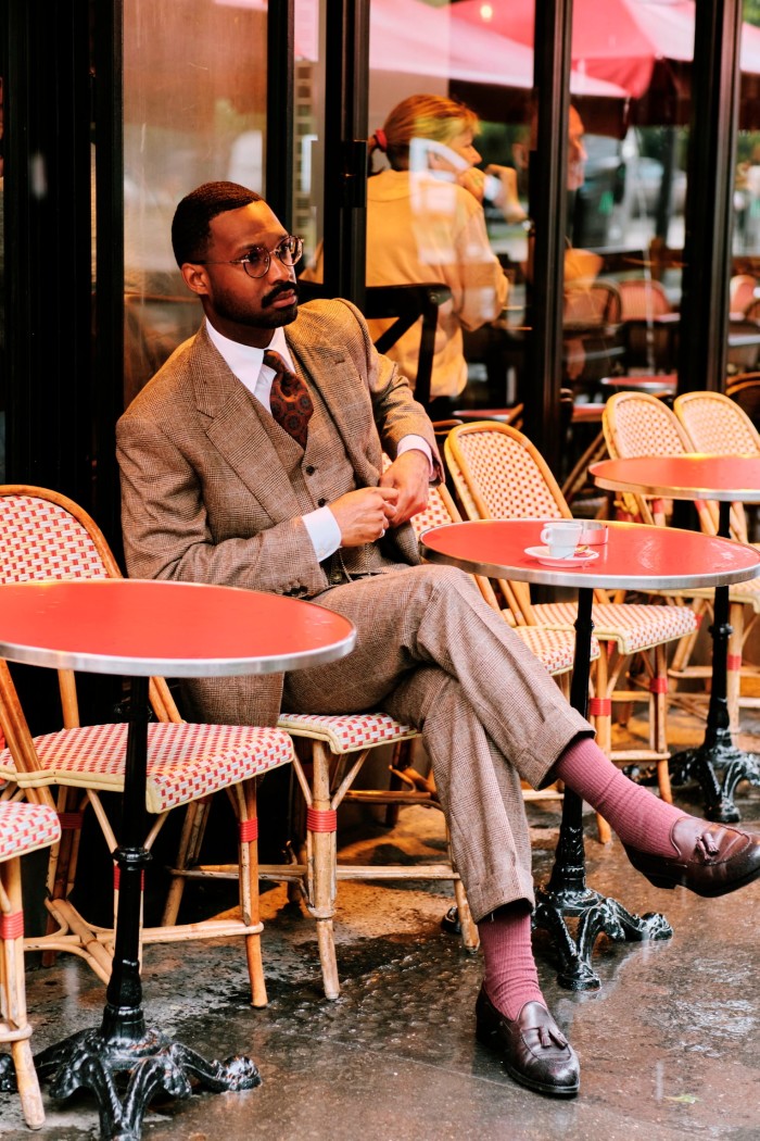 A man in a beige check three-piece suit sits at a French cafe table