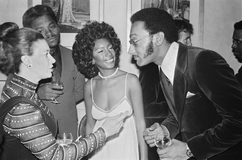 In a black and white photograph, a woman is shown speaking to a man in glasses and a suit as two other people look on.