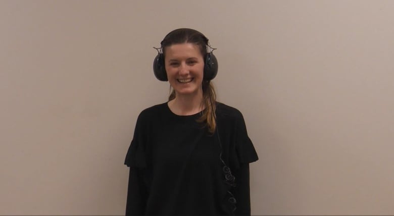 A woman wearing large headphones stands against a beige wall smiling and blushing