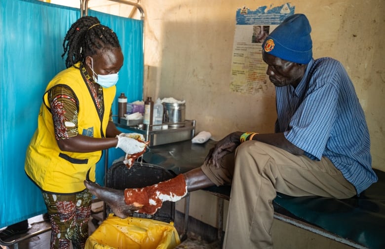 A woman helps a man seated on an examination table with a bandage on his leg. 