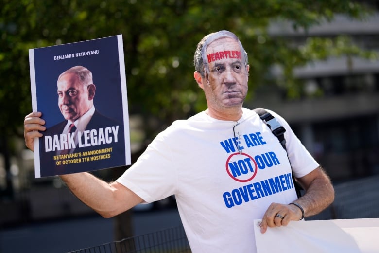 A demonstrator wears a mask and a tshirt in an outdoor setting. He's holding up a sign of another man's image, which says 'Dark Legacy.'