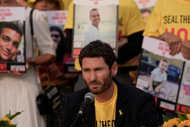 A bearded man is shown speaking into a microphone as signs are shown behind him depicting the images of assorted individuals.