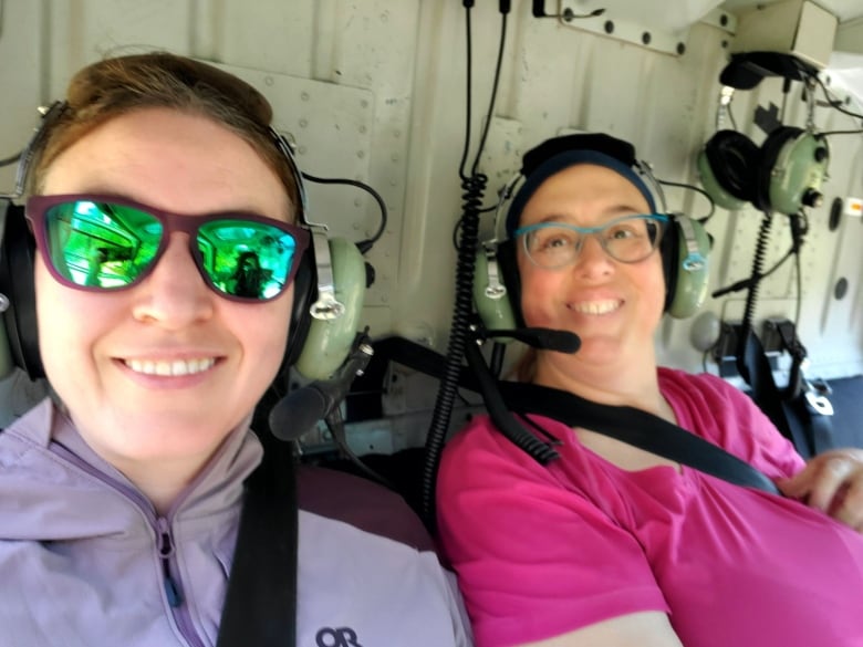 Two women smiling and wearing headphones while strapped into a helicopter.