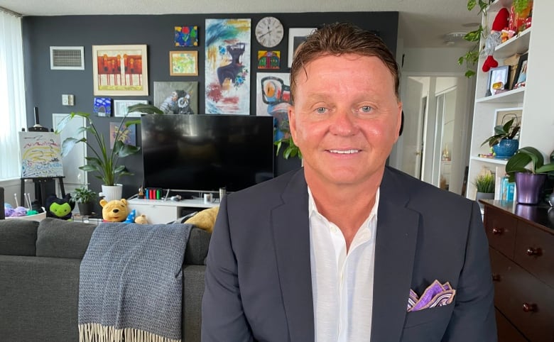 Steve Moran, wearing a suit jacket, sits in a living room with colourfuul artwork on the wall behind him. 