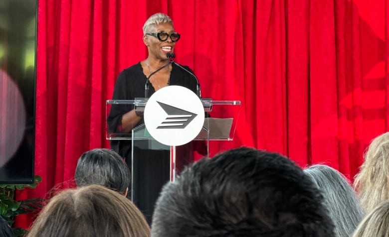 Maxine Bailey stands in front of a podium, with a red curtain backdrop delivering a speech about Norman Jewison