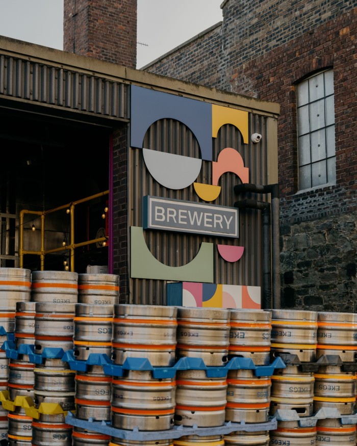 Piles of beer kegs in front of a sign that says “Brewery” and colourful cut-out shapes on a corrugated wall at Moonwake Beer Co