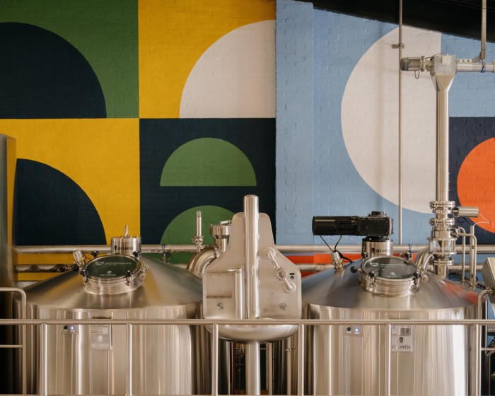 Stainless-steel brewing equipment in front of a wall covered in yellow, green and blue squares on which sit dark, white and green semi-circles