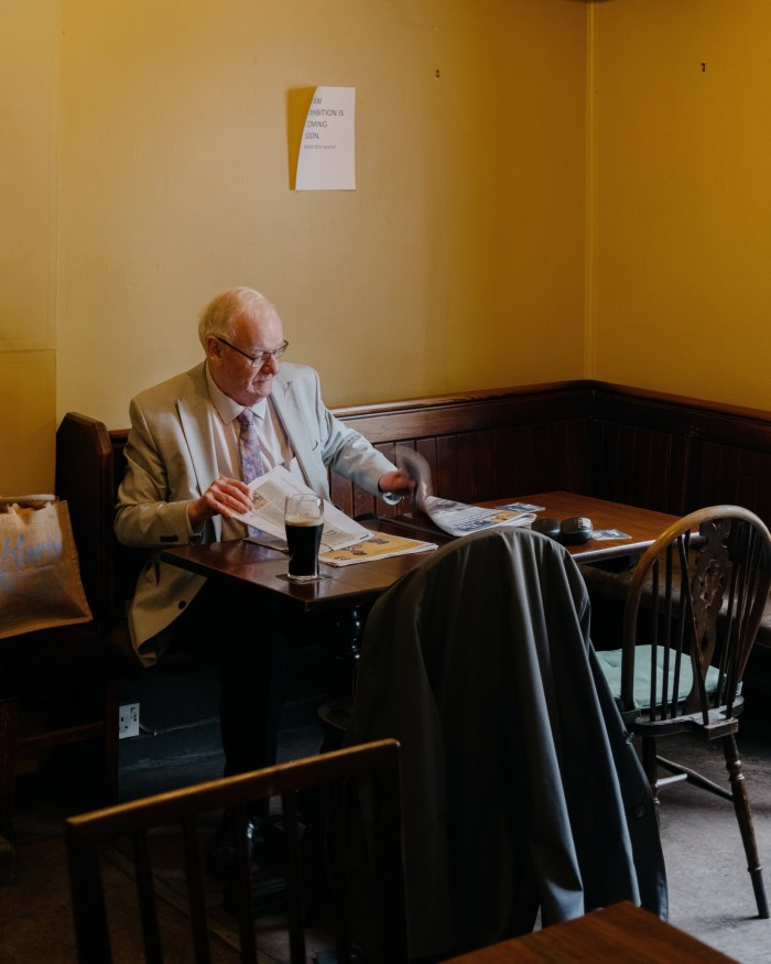 An elderly man in a suit reading a newspaper while drinking a pint of stout at a table in The ‘Oxford’