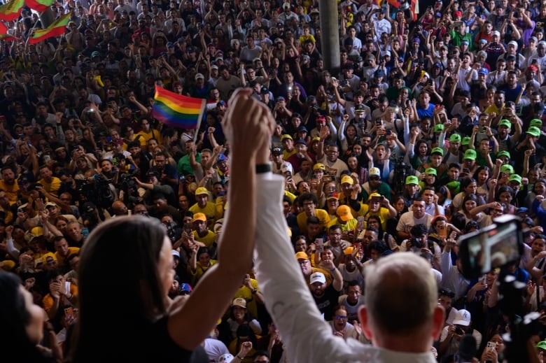 A man and a woman are seen from behind with arms raised above their heads, looking out at a crowd of faces that appears to number in the thousands.