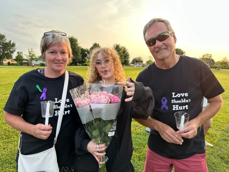 Heather Trevithick and Joe Quigg stand with Broadfoot's friend Tavia Lapier, middle, wearing T-shirts that say "Love shouldn't hurt" next to a purple ribbon, which is a symbol of opposition against IPV.