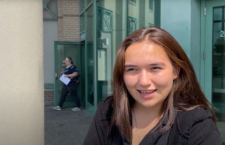 A young woman with long, brown hair speaks outside the glass doors of a building.