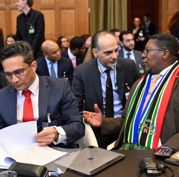 Two men in suits and one man wearing a sash in the colours of the South African flag over his suit all seated in a courtroom