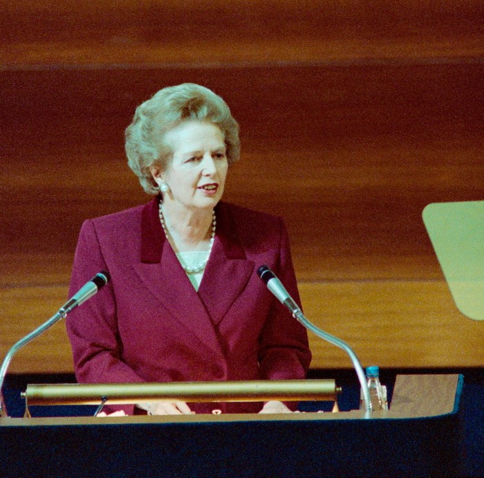 Margaret Thatcher wearing a mauve coloured suit at a podium behind two microphones