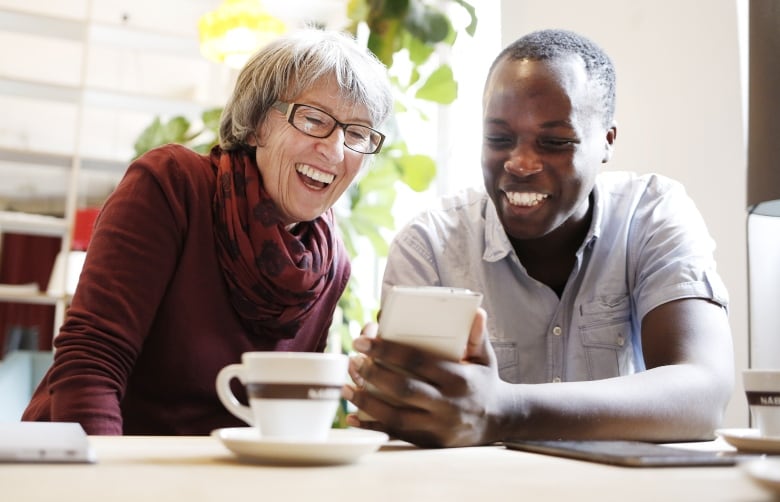 An older woman sits next to a young man at a table as they both smile broadly while watching something on his smartphone.