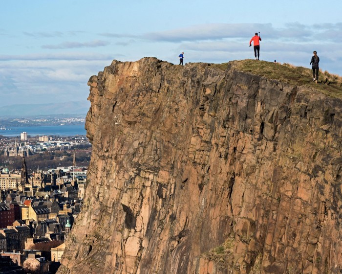 The craggy sheer cliff of Arthur’s Seat in Edinburgh, with three runners at the top