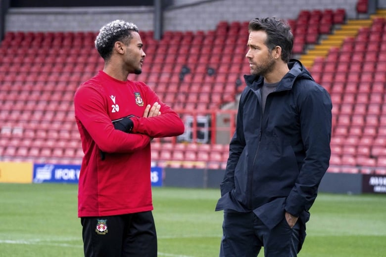 A player in a red football kit speaks to a man wearing a navy blue sports jacket on a football pitch.