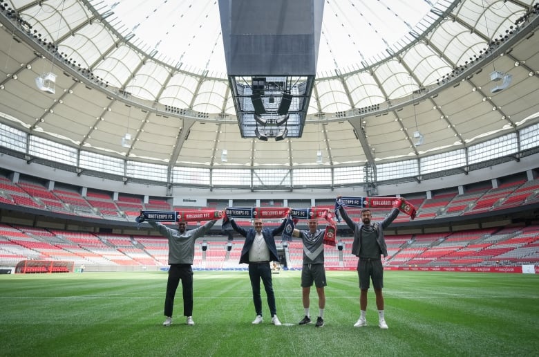 Four men stand on a soccer pitch with scarves belonging to their respective teams displayed over their heads.