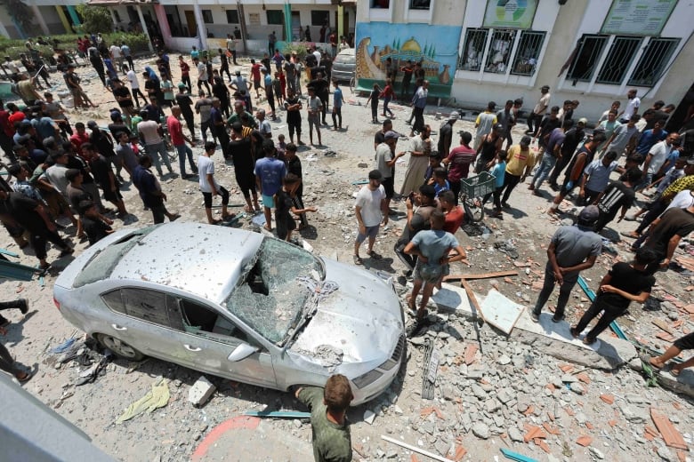 People inspect a street outside a building that is covered in debris with a damaged car.