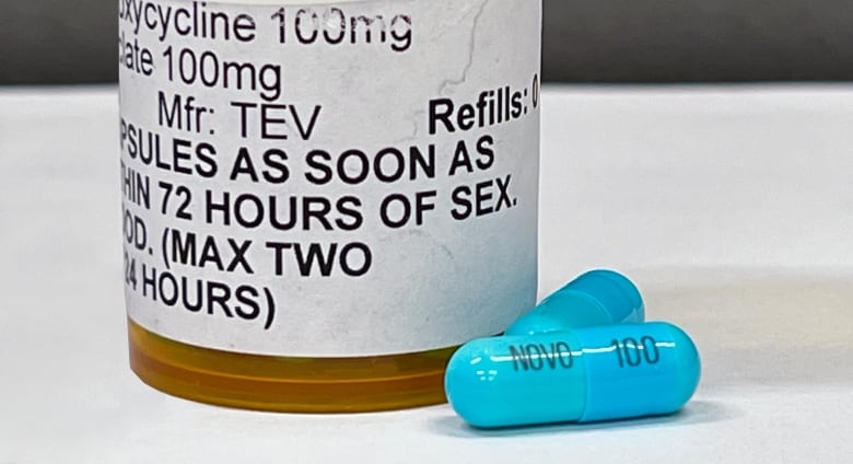 Two blue capsules on a white surface next to an orange prescription bottle with a white label and instruction printed in black on it. 