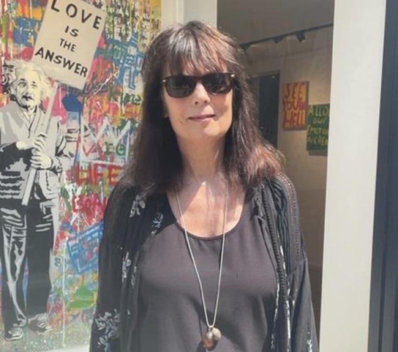 A woman wearing mostly black with sunglasses and a long necklace smiles for a photo. A graffiti art mural is in the background featuring an image of Albert Einstein holding a sign that reads "Love is the answer."