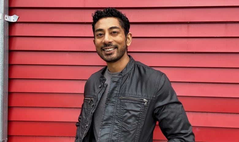 A man smiles for a portrait with his hands in the pockets of his leather jacket in front of the red wooden exterior of the building behind him.