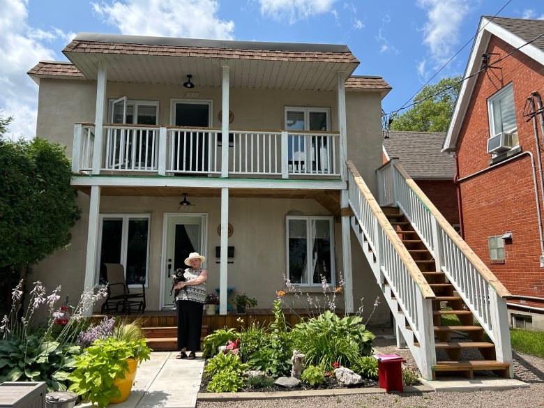 A woman holding a black dog in her arms stands in front of a beige duplex. 