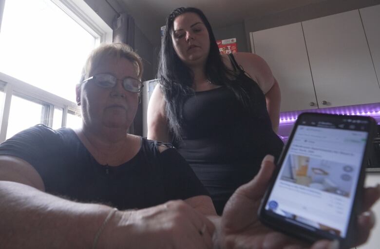 A woman holds a cellphone in her hand pointed towards the camera, while another woman stands in the background looking on.