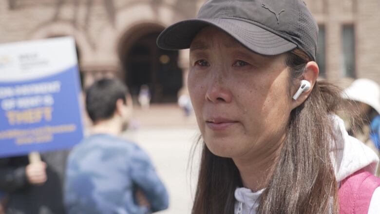 A woman in a ball cap and with a distraught look on her face stands outdoors in a crowd.