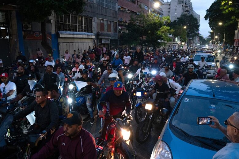 A throng of motorbikes drive on an urban street at night.