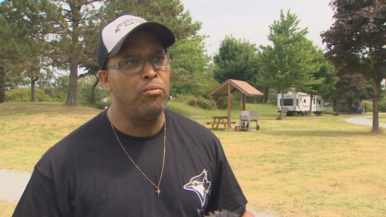 Man wearing a black t-shirt and a ball cap stands in a park.