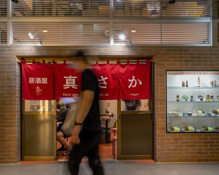 The red-brick facade of Izakaya Masaka, with a window showing the dishes on offer and red cloth banners hanging over the doors. An out-of-focus man is walking by