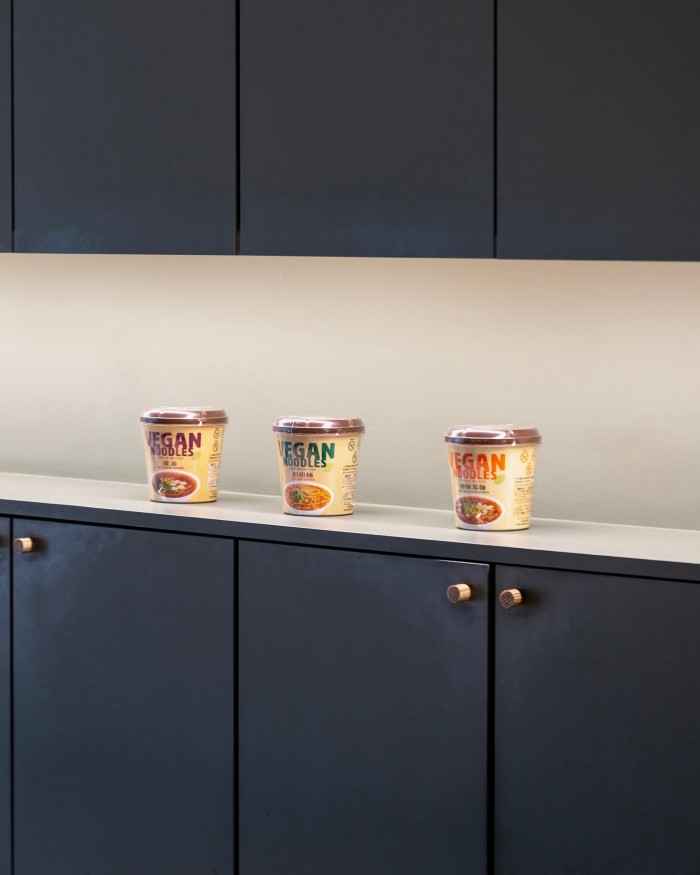 Three tubs of vegan noodles on a lit shelf between black-wood cupboards at Vegan Bistro Jangara 