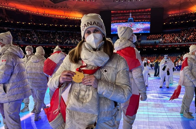 A woman dressed in Team Canada gear and wearing a mask poses with a gold medal.