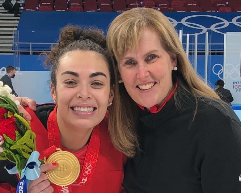 Two women, one holding a gold medal, smile together on the ice.