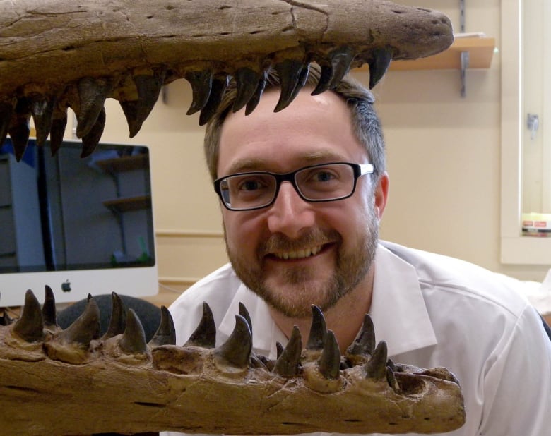 A man with glasses smiles as he peers through the open, tooth-laden mouth of a fossil. 