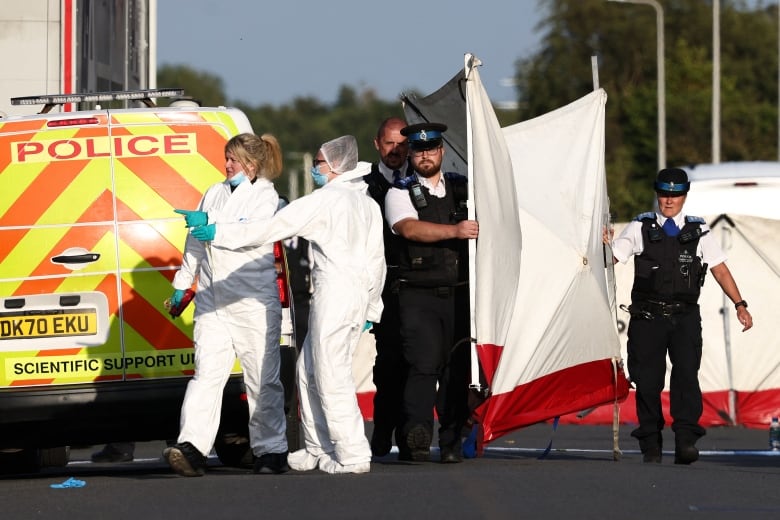 Two people in white suits and latex gloves stand near a police van with the words "scientific support" on it. Three nearby police carry a barrier.