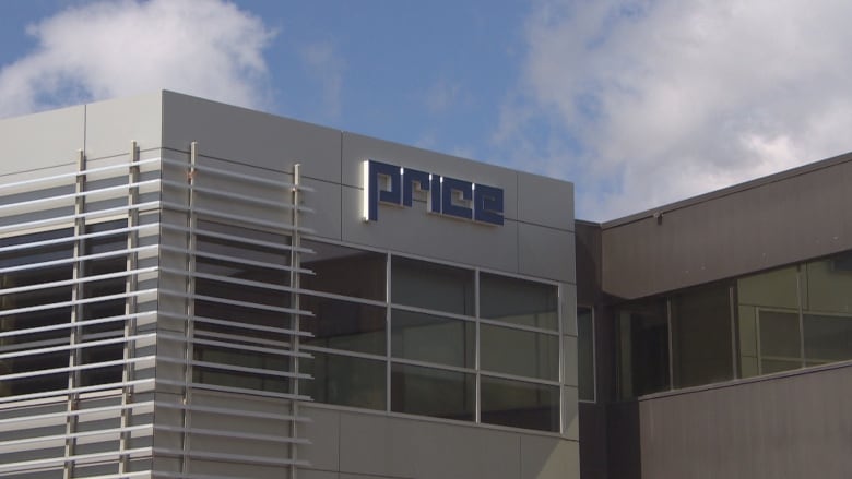 A grey building is seen with window panels and a sign with the name "Price" near the top, a blue sky and clouds in the background.